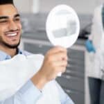 Happy Handsome Arab Man Looking To Mirror After Teeth Treatment In Modern Clinic, Middle Eastern Male Patient Sitting In Chair In Stomatological Cabinet And Enjoying His New Smile, Closeup